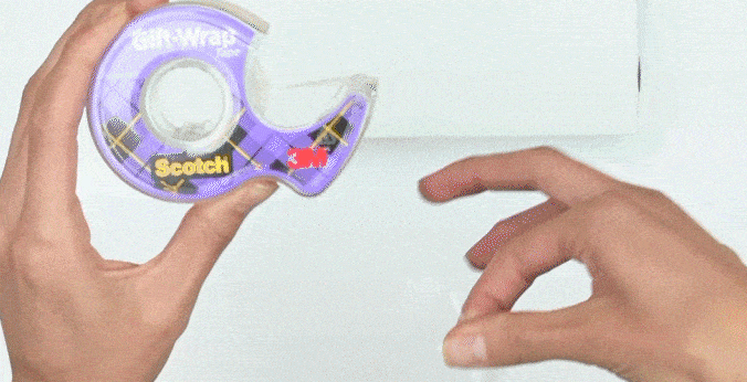 Manos de mujer envolviendo una caja con papel y pegando los extremos con Cinta de Regalo Scotch®.
